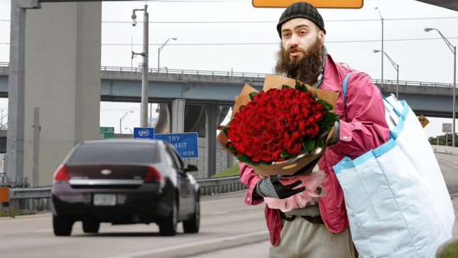 Man with tan skin and brown hair with a scraggly black beard and black beanie standing on the side of a highway. He's holding a bouquet of red roses, wearing a faded red shirt and has a white bag on his side. There is a black car passing him. It is during the day, slightly overcast.