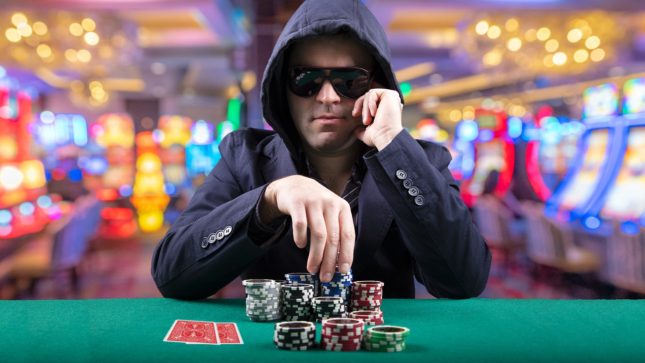 White guy in a black sweatshirt with black, thick sunglasses. He has one hand resting against his face that has an angry expression. His other hand is limply hanging over a green poker table. He has a drink in front of him and cards. There are lights and games in the background.