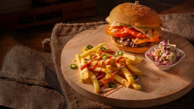 Fancy cheese burger with fries that have ketchup and jalapenos sprinkled on them. Served on a round wooden board over a brown table cloth on a dark table.