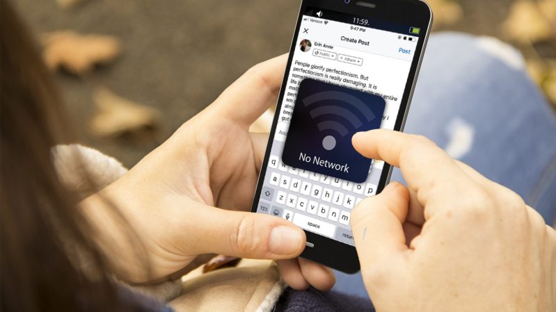 White woman scrolling on phone with a typed out status of her oversharing and a black button that says "No Internet". She is wearing light washed, ripped jeans and you can see the ground; there are leaves. She has black hair.