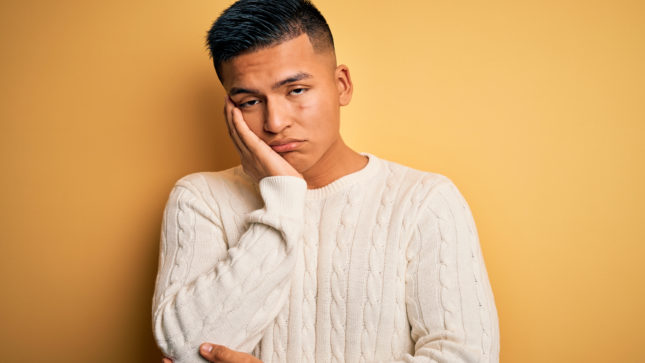 Man with tan skin and acne in a white knit sweater with his hand on his face. He looks sad. There is a yellow background. His hair is black and short and his eyes are brown and he looks sad.