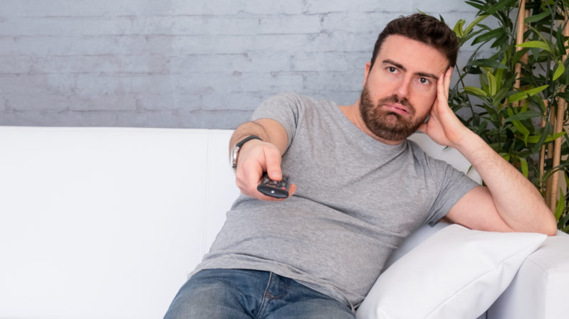White guy with brown hair and brown eyes looking annoyed with his arm against the side of his face. He's in a gray shirt and jeans sitting on a white couch with a gray background and a plant behind him. He is holding a remote out.