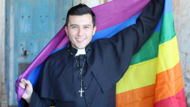 A pale man in his mid-20s with black hair and brown eyes wearing a priest suit black and white, holding a rainbow flag against a blue background while smiling.