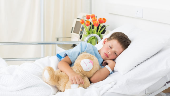 White boy in hospital bed with sandy blonde hair, his eyes are closed and he's clutching a teddy bear. There are orange/yellow roses behind him and a monitor and tan drapes.