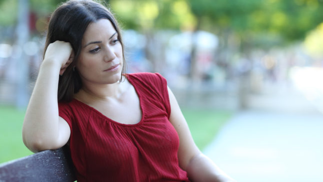 White woman in red v-neck shirt looking sad with her fist against her head. She has brown hair and brown eyes and is sitting in a park on a bench.