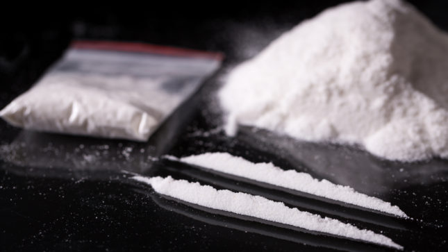 Two lines of cocaine on a black table next to a bag of cocaine and a white mound of cocaine.