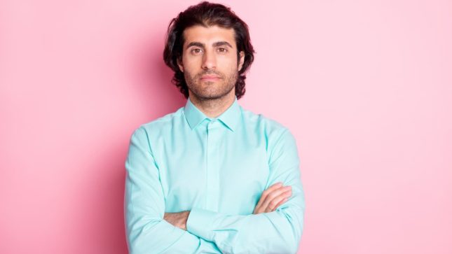 Man with darker tan skin crossing his arms. He has thick eyebrows and shoulder length black hair and brown eyes. He has a neutral facial expression and is crossing his arms. He is wearing a turquoise shirt and there is a pink background.