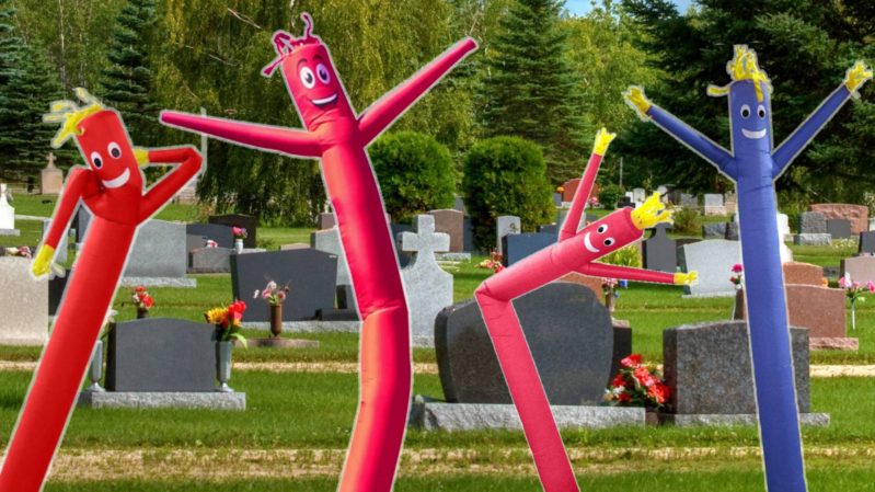 Red and blue inflatable tube men in front of tombstones on greenish-yellow grass.